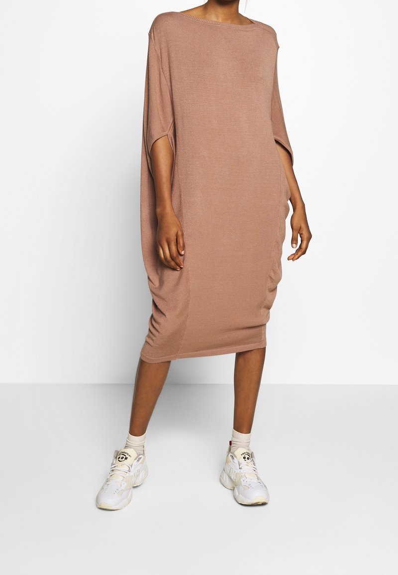 Robe marron tricotée surdimensionnée à manches courtes, avec fronces latérales et silhouette décontractée, associée à des baskets blanches et des chaussettes beiges.