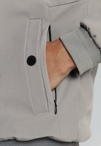 Grijze jas met een ritszak, zachte stof, gebreide manchetten en een afgeronde onderkant. Voorzien van een zwart drukknopdetail.