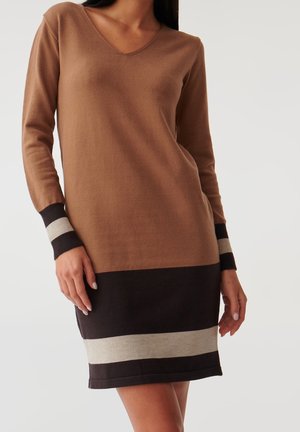 Kobieta w brązowej, długorękawowej, dzianinowej sukience z dekoltem w kształcie litery V oraz czarnymi i beżowymi poziomymi paskami na mankietach i dole sukienki, stojąca na jednolitym tle.