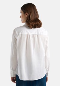 Camicia bianca a maniche lunghe, in cotone, con polsini abbottonati, dettaglio arricciato sul retro, orlo curvo, indossata con una gonna blu plissettata.