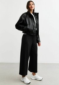 Zwarte leren cropped jasje met drukknoopsluitingen, gecombineerd met zwarte broek met wijde pijpen en wit sportief schoeisel.