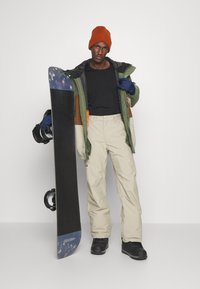 Snowboarder con pantalones beige, camiseta negra y chaqueta verde-marrón, sosteniendo una tabla de snowboard oscura con punta azul. Un gorro naranja y botas negras completan el atuendo.