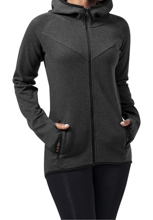 Vrouw met een donkergrijze hoodie met rits en duimgaten, handen in de zakken, gecombineerd met zwarte leggings, zichtbaar van nek tot heupen.