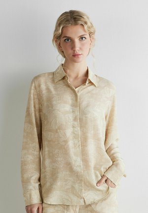 Blonde Frau trägt beige Hemd mit dezentem Blattmuster, steht vor schlichter heller Wand, Hand in der Tasche, schaut in die Kamera.