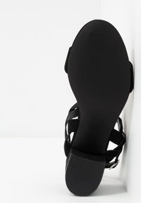 Sandale à talon en suède noir avec une semelle texturée, arrière ouvert et un design de lanières croisées. Elle dispose d'un talon plat et d'une bride de cheville ajustable.