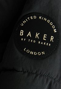 Pezzo di stoffa nera a forma circolare, con lettering dorato in rilievo: "BAKER by TED BAKER," "UNITED KINGDOM," "LONDON," e dettaglio di cucitura.