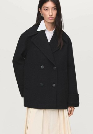 Black double-breasted coat made of heavy fabric, featuring large notched lapels, two front pockets, and black buttons. Styled over a white shirt.