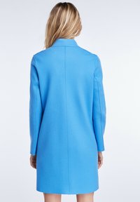 Manteau en laine bleue avec une coupe ajustée, manches longues et coupe droite. Texture lisse et coutures peu visibles à l'arrière.