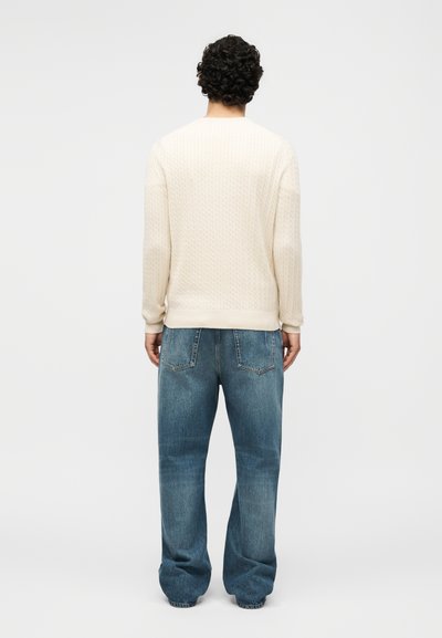 Άνδρας με σγουρά μαλλιά που φοράει ένα κρεμ πουλόβερ με πλέξη καλώδιο και άνετο μπλε τζιν, στέκεται γυρισμένος με την πλάτη σε ένα απλό λευκό φόντο.