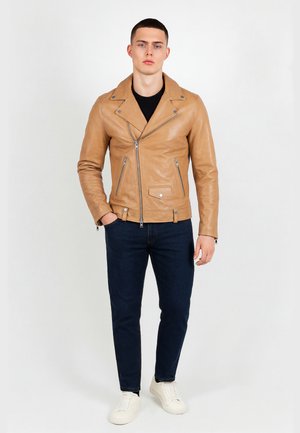Młody mężczyzna stoi, ubrany w jasnobrązową skórzaną kurtkę motocyklową, czarną koszulę, ciemnoniebieskie dżinsy i białe trampki na białym tle.