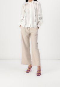 Valkoinen pitkähihaisella puserolla, jossa on pitsiyksityiskohtia, yhdistettynä beigekuituihin leveisiin housuihin ja purppuranvärisiin nauhakorkokenkiin. Silkkinen pinta ja rento malli.