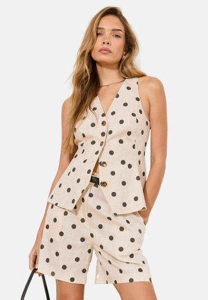 Frau trägt beige ärmellose Weste mit Punkten und passende hoch taillierte Shorts, hält eine schwarze Handtasche, mit lockerem welligem Haar.