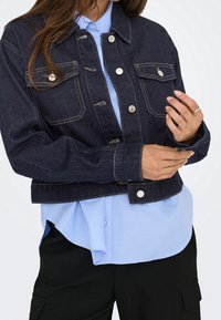 Personne portant une veste en jean bleu foncé sur une chemise bleu clair et un pantalon noir, ajustant la manche de la veste avec ses mains.