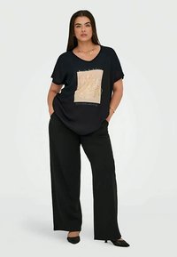 Naine, kellel on seljas mustad laiad püksid ja must T-särk, millel on beež graafika, seisab valge tausta ees, käed taskutes.