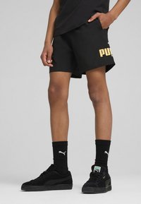 Pantalones cortos negros con el logotipo dorado de "PUMA", hechos de algodón y con un corte relajado. Combinados con zapatillas negras y calcetines negros de canalé.