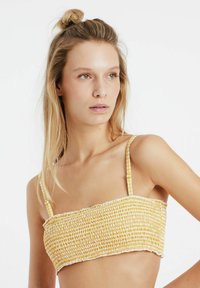 Geel smocktop met korte mouwen en dunne bandjes, gemaakt van een textuur met elastisch materiaal en een zachte, gerafelde zoom. Eenvoudig ontwerp en neutrale achtergrond.