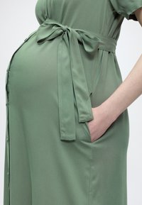 Έγκυος φοράει πράσινο φόρεμα με δεμένη ζώνη στη μέση και έχει το χέρι της μέσα σε πλαϊνή τσέπη.