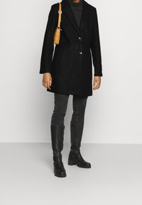 Manteau noir en mélange de laine avec un col cranté, deux boutons et des poches latérales, associé à un jean skinny gris foncé et des bottes noires montantes jusqu'aux genoux.