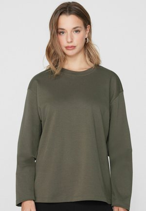 Žena se světle hnědými vlasy po ramena, která nosí stříbrné kruhové náušnice a olivově zelené tričko s dlouhým rukávem a kulatým výstřihem, čelem k fotoaparátu.