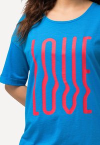 Mujer vestida con una camisa de color azul brillante con letras rojas en forma de ondas que dicen "AMOR" en el frente, mostrada desde el cuello hasta la cadera.