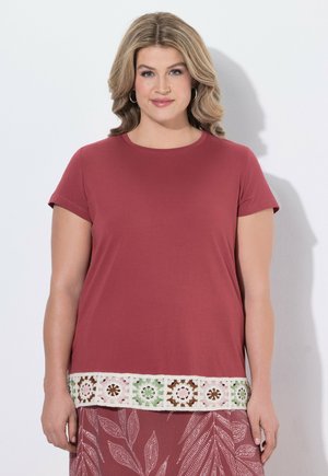 Frau mit blonden Haaren trägt Creolen, ein kurzärmeliges rosafarbenes Oberteil mit floraler Häkelbordüre und einen passenden gemusterten Rock.