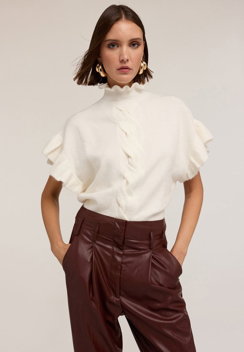 Suéter corto de punto blanco con bordes ondulados y un cuello alto, que presenta un diseño de trenzas centrales, combinado con pantalones de imitación de cuero burdeos.
