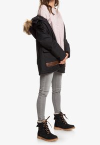 Parka noire avec capuche bordée de fourrure, accents beige, portée sur une chemise rayée rose. Jean slim gris et bottes noires avec doublure en fourrure et détails en lacets.