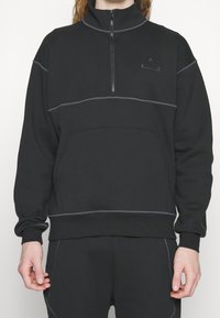 Sudadera negra con cremallera hasta el cuarto, costuras grises, bolsillo frontal y detalle sutil de logo en el pecho. Hecha de un material suave.