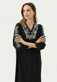 Schwarzes Kleid mit bestickten weißen Blumenmustern entlang des Ausschnitts und der Ärmel; mit V-Ausschnitt und dreiviertellangen Puffärmeln.
