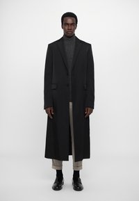 Cămașă lungă neagră cu un design structurat și închidere cu un singur nasture, purtată peste un pulover gri cu guler înalt, asociată cu pantaloni bej și pantofi negri.