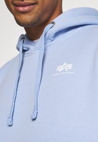 Homme portant un sweat à capuche Alpha Industries bleu clair avec cordons et un sourire partiel visible.