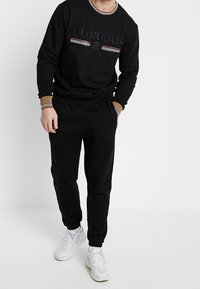 Felpa nera con la scritta "GLORIOUS" e accenti rigati, abbinata a pantaloni della tuta coordinati e scarpe da ginnastica bianche. Tessuto morbido e casual.