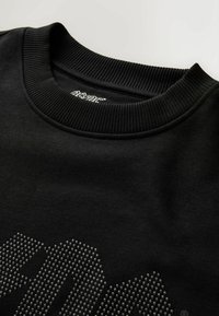 Fekete pulóver bordázott kerek nyakkal, elülső része emelt pöttyös mintával és diszkrét logóval a gallér közelében.