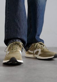 Olivengrønne atletiske sko med en blanding af ruskind og mesh, hvide accenter og en sort gummisål, bæres med denim jeans.