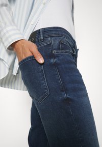 Main dans la poche arrière d'un jean bleu foncé porté avec une chemise blanche rentrée et une chemise à rayures bleu clair boutonnée.