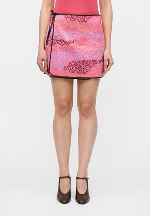 Ženska nosi roza mini ovijalno krilo z grobo teksturo in abstraktnimi vzorci, kombinirano s črnimi čevlji Mary Jane, na belem ozadju.