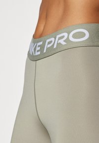 Oliven grønne sportsleggings med en bred elastisk talje, der har "NIKE PRO" i hvid. Glat tekstur med en tætsiddende pasform.