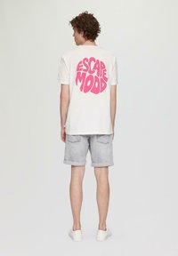 Weißes Baumwoll-T-Shirt mit großem pinkem "ESCAPE MOOD"-Grafik auf dem Rücken, kombiniert mit hellgrauen Shorts und weißen Sneakers.