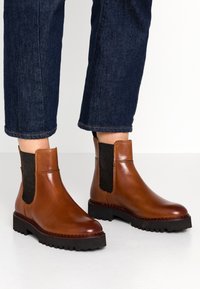 Bottines en cuir marron avec des panneaux latéraux élastiques, semelles en caoutchouc noir épaisses et détails de couture. Portées avec un jean en denim foncé.