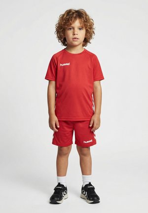Enfant aux cheveux bouclés debout, portant un maillot de sport rouge et un short, des chaussettes blanches et des chaussures de sport noires sur fond blanc.