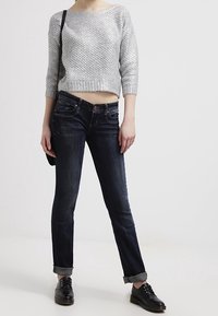 Suéter gris de punto con mangas tres cuartos, combinado con jeans oscuros de corte ajustado y zapatos negros de cordones. Look casual con los puños arremangados.