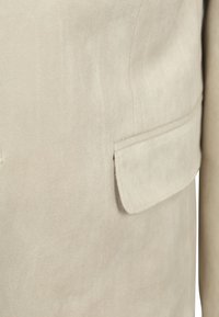 Beige suede jasje met een borstzak aan de linkerkant en een gladde textuur. De stof heeft een zachte, licht reflecterende uitstraling.