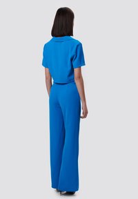 Mono azul con mangas cortas y cuello, hecho de tela suave. Cuenta con pantalones de pierna ancha y una cintura ajustada. Visto desde la parte de atrás.