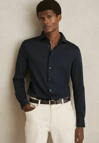 Cămașă albastru închis cu mânecă lungă și guler à la button-down, cu nasturi în față, purtată cu pantaloni crem și o curea texturată maro.