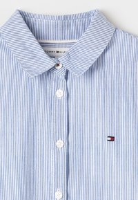 Camicia a righe blu e bianche con bottoni frontali e colletto ampio, caratterizzata da bottoni bianchi e un piccolo logo rosso, bianco e blu sul petto.