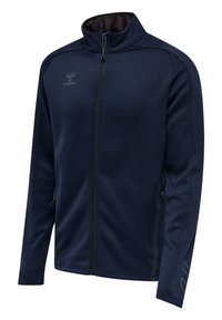 Veste zippée bleu marine en matière lisse et extensible. Elle présente un col haut, des poches latérales zippées et un logo discret sur la poitrine.