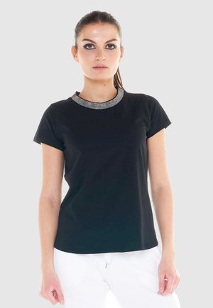 Femme portant un haut noir à manches courtes avec un col orné de bijoux et un pantalon blanc, debout devant un fond clair uni.