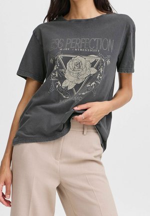 Vrouw draagt een donkergrijs T-shirt met grafisch rozen- en driehoekmotief, beige broek en zilveren ringen, staand tegen een effen achtergrond.
