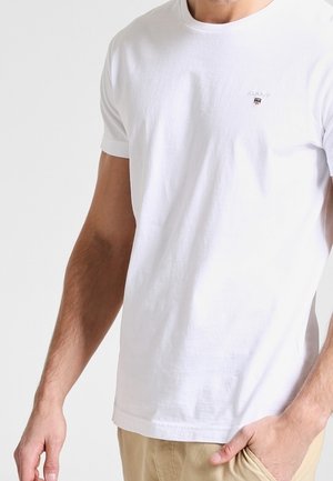 Homme portant un t-shirt blanc uni à manches courtes avec un petit logo GANT sur la poitrine, pantalon beige, main dans la poche, sur fond blanc.