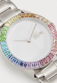 Reloj plateado con una esfera blanca, adornado con un bisel de gemas rectangulares de colores múltiples. Pulsera de acero inoxidable, con manecillas de horas y minutos minimalistas.